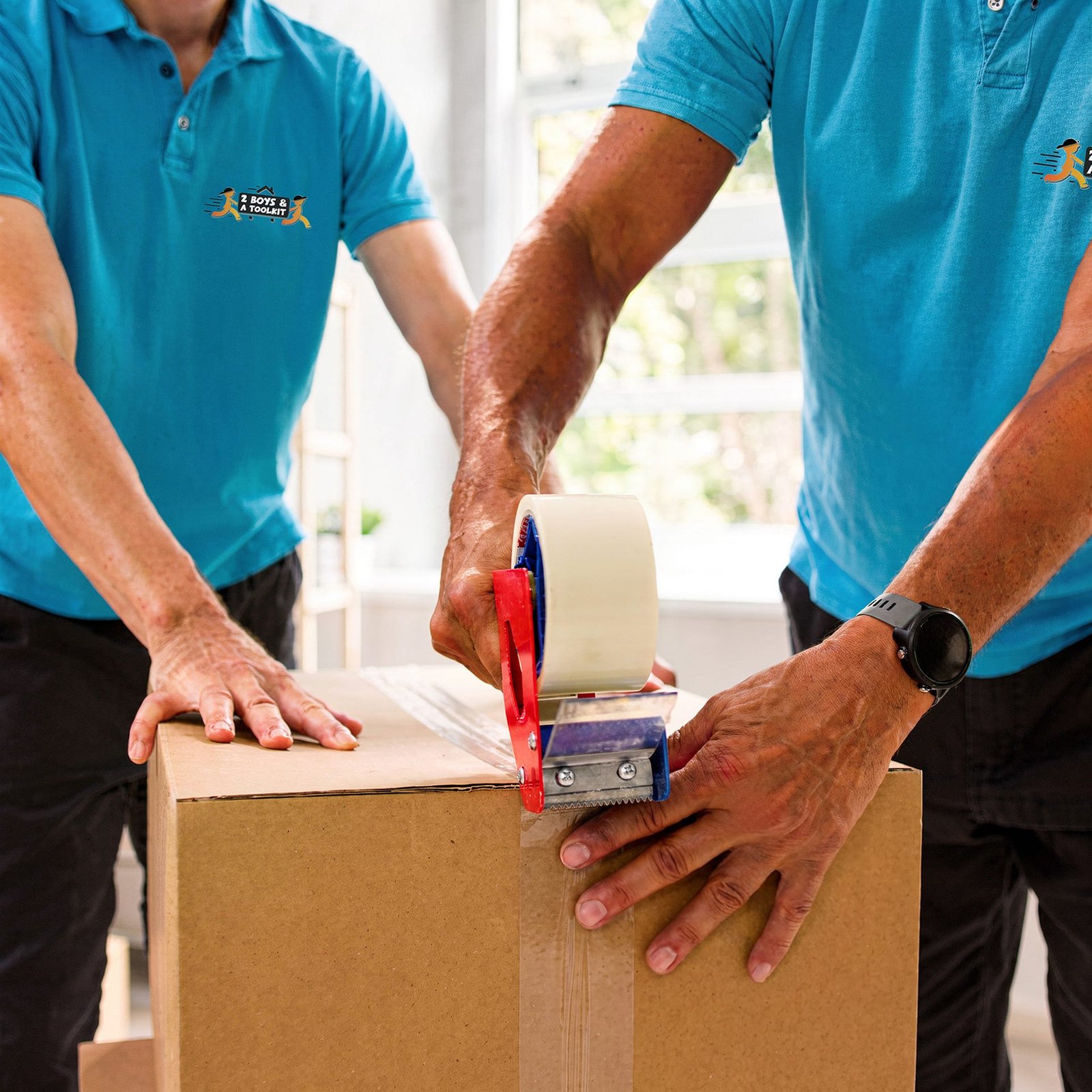 Two 2 Boys & A Toolkit movers in teal shirts sealing a cardboard moving box with packing tape