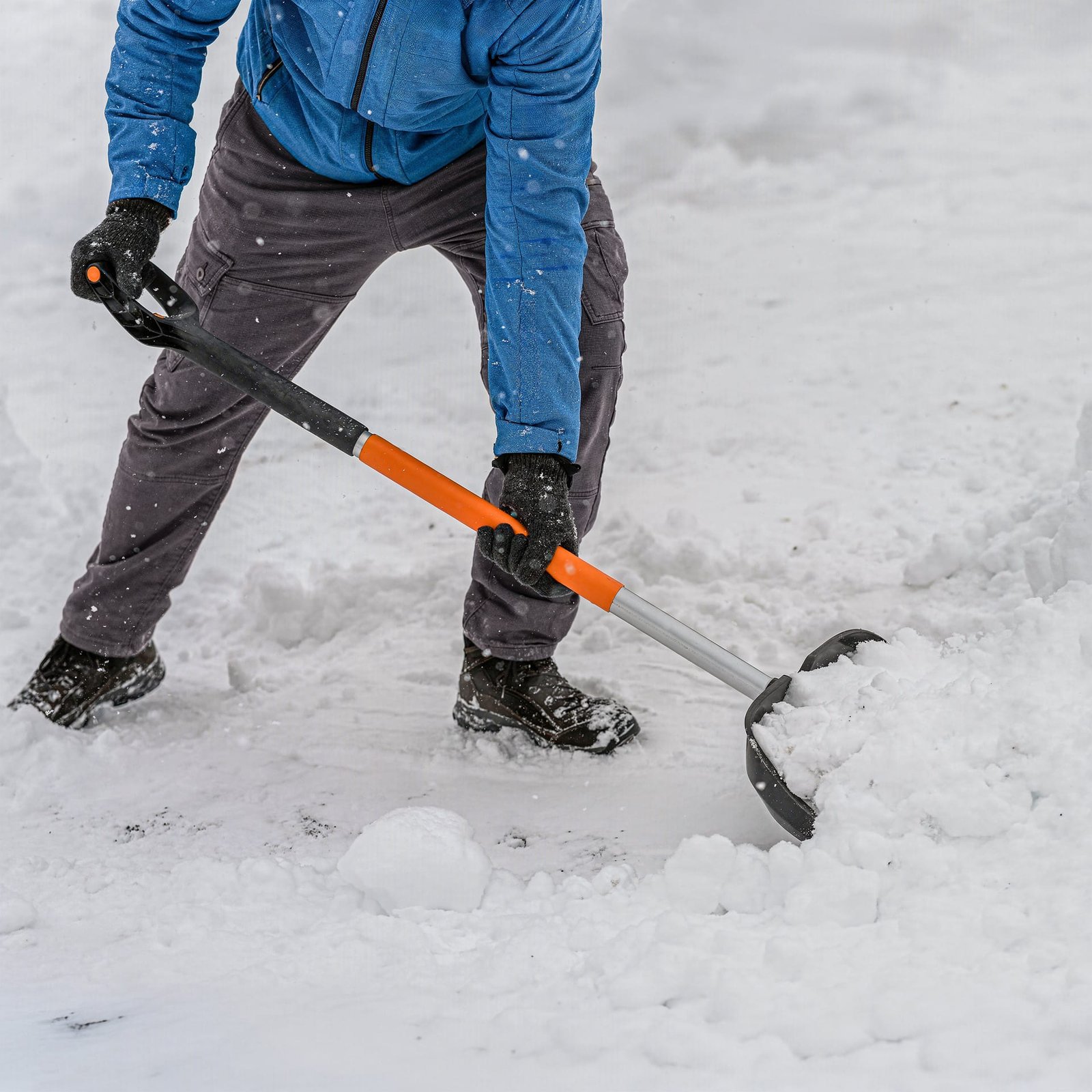 Person in winter clothing using an orange and grey shovel to clear snow from the ground