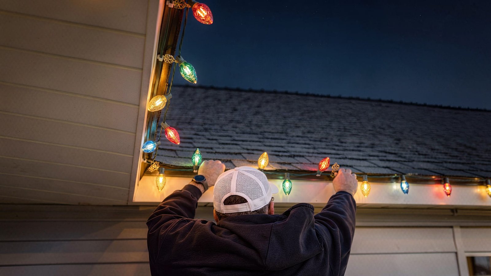 Man reaching up to hang coloured Christmas lights along the roofline of a house at dusk