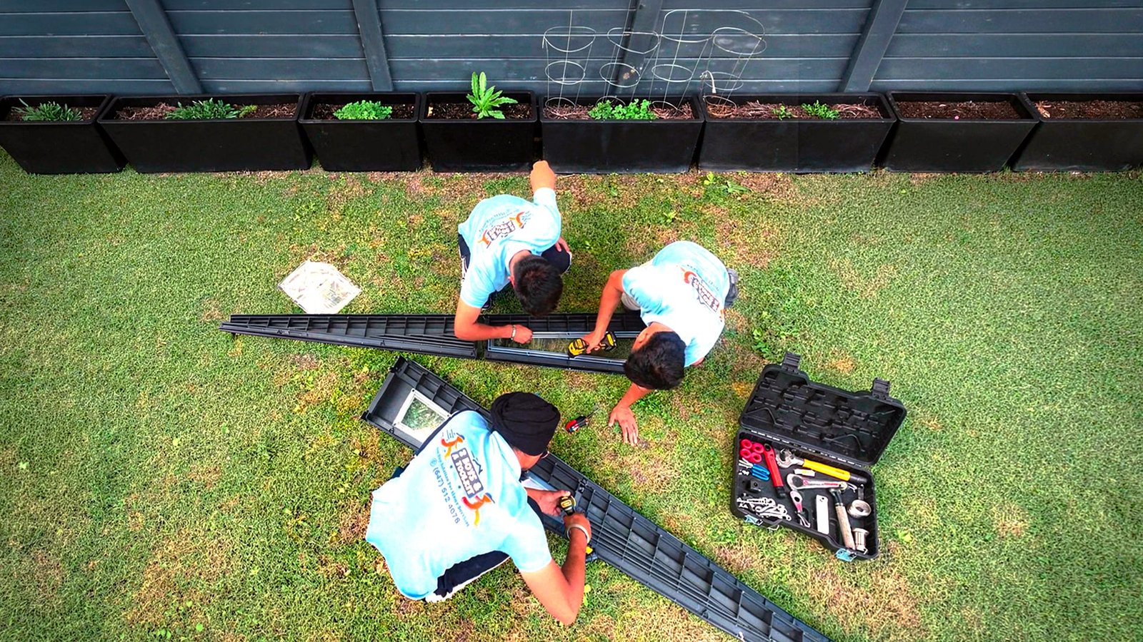 Overhead view of three workers assembling raised garden bed frames on a lawn near planter boxes, using power drills and a toolbox of tools