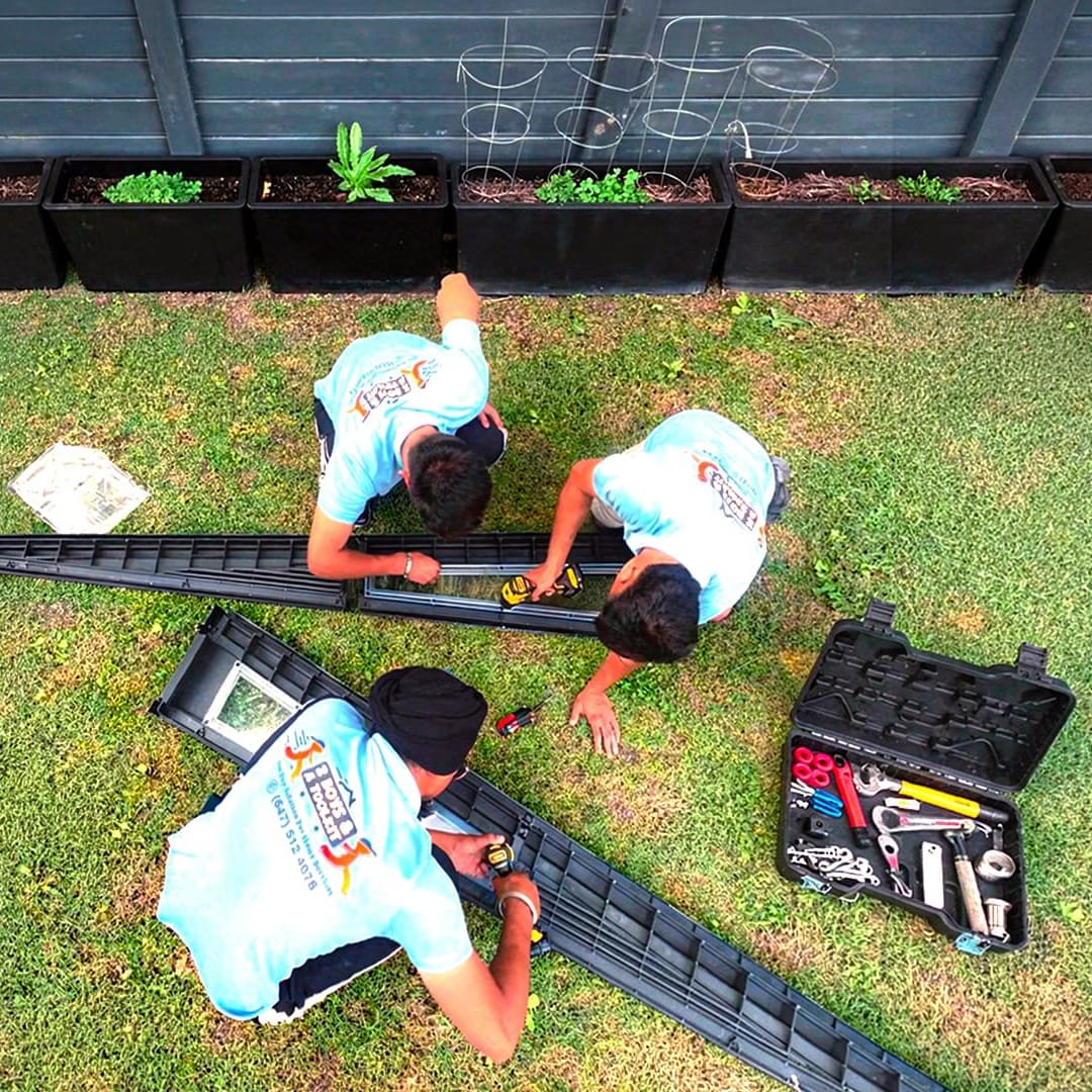 Overhead view of three workers in matching shirts assembling raised garden bed frames on grass, with a toolbox and drill nearby