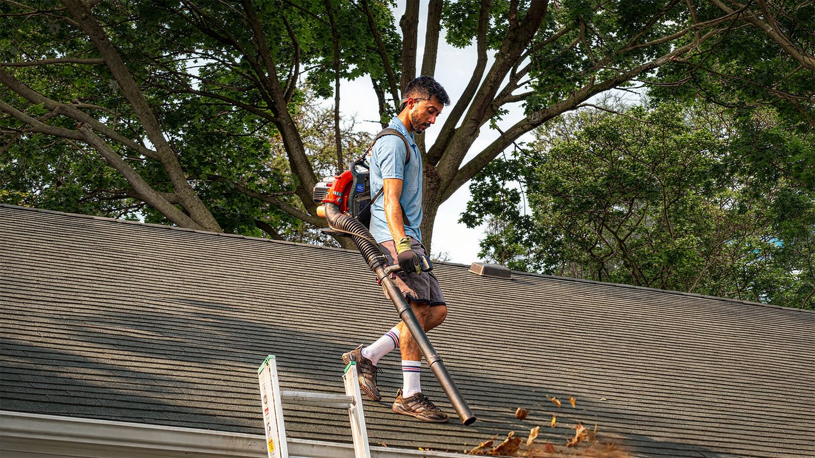 Worker on a rooftop using a backpack leaf blower to clear debris from the roof and gutters