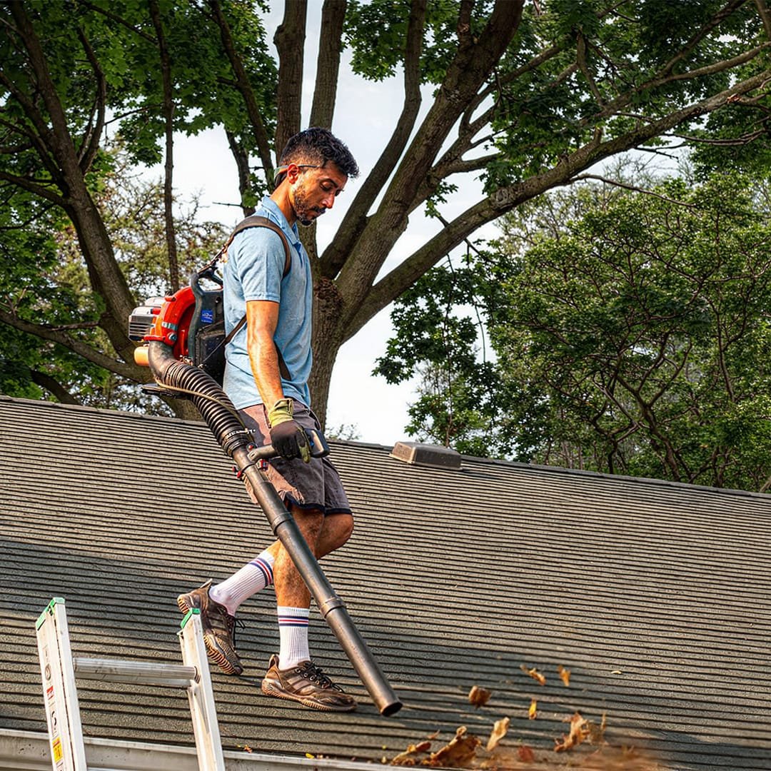 Worker on a rooftop using a backpack leaf blower to clear debris from shingles near a ladder