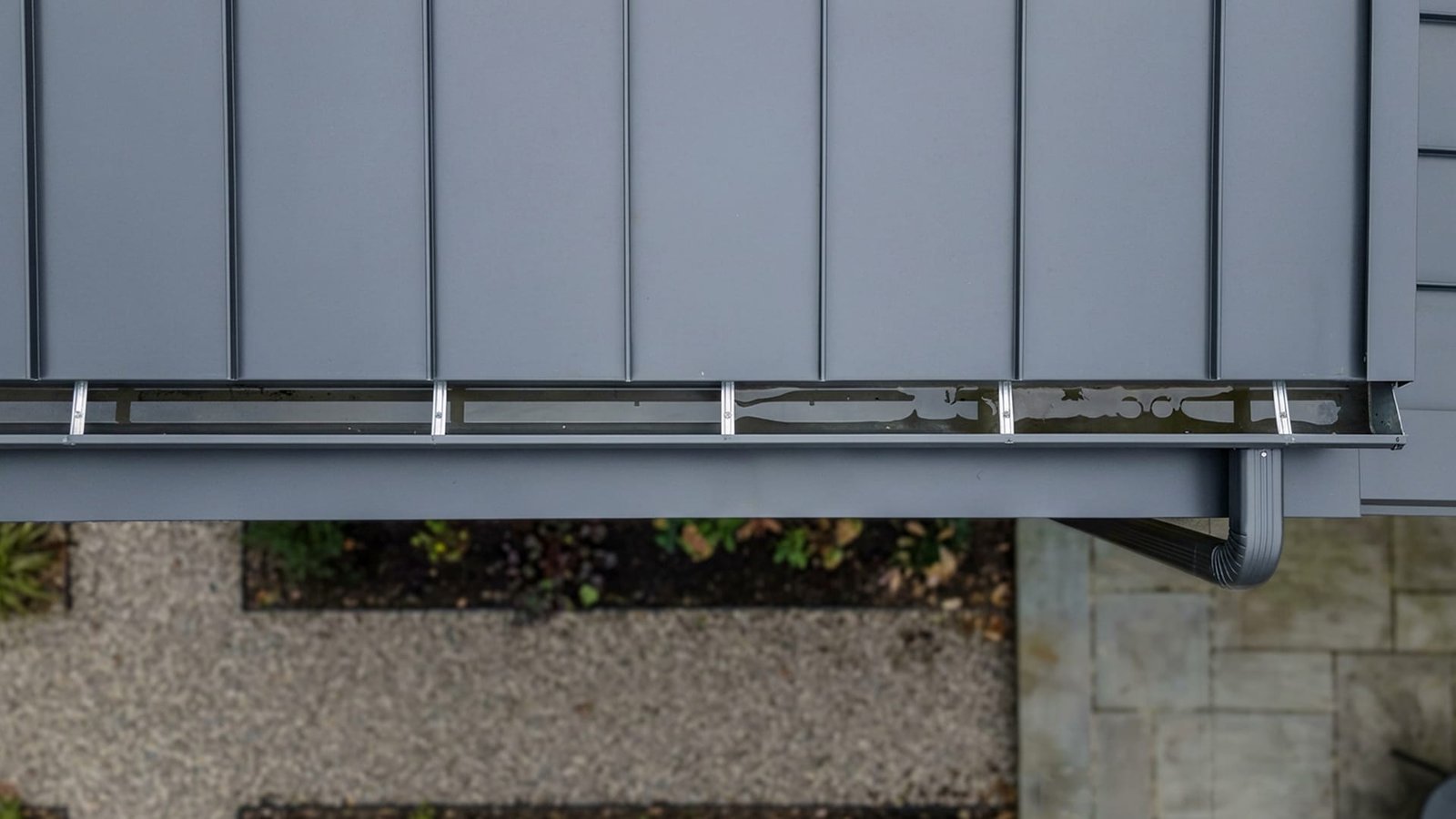 Close-up of a grey standing-seam metal roof edge with integrated gutter and downspout above a gravel path and garden bed