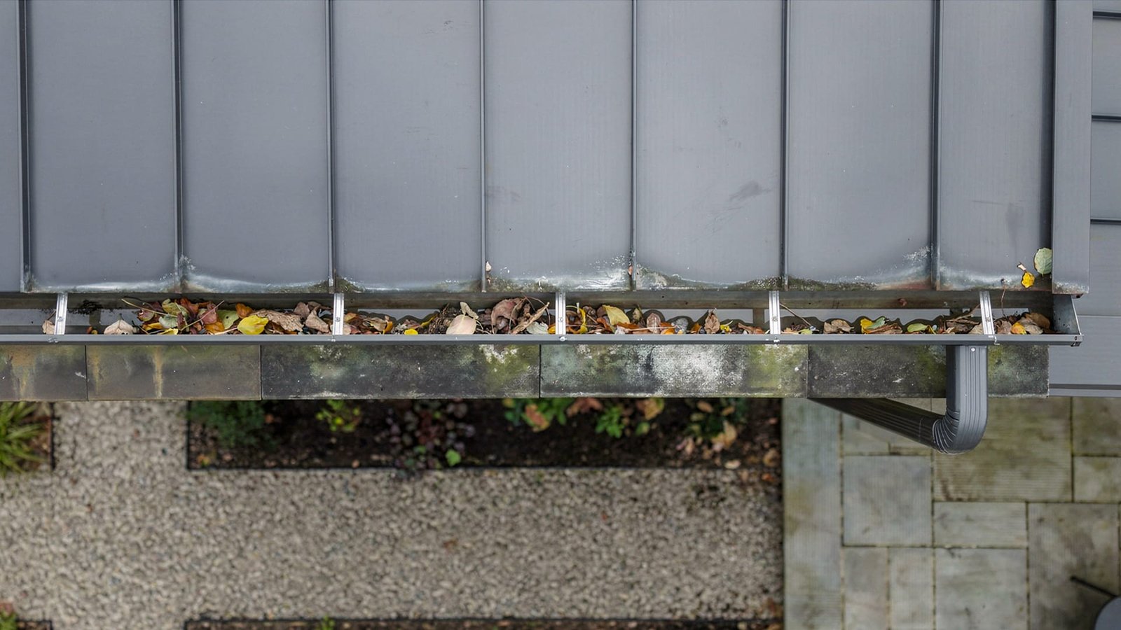 Metal roof gutter clogged with dry leaves above a paved and gravel walkway