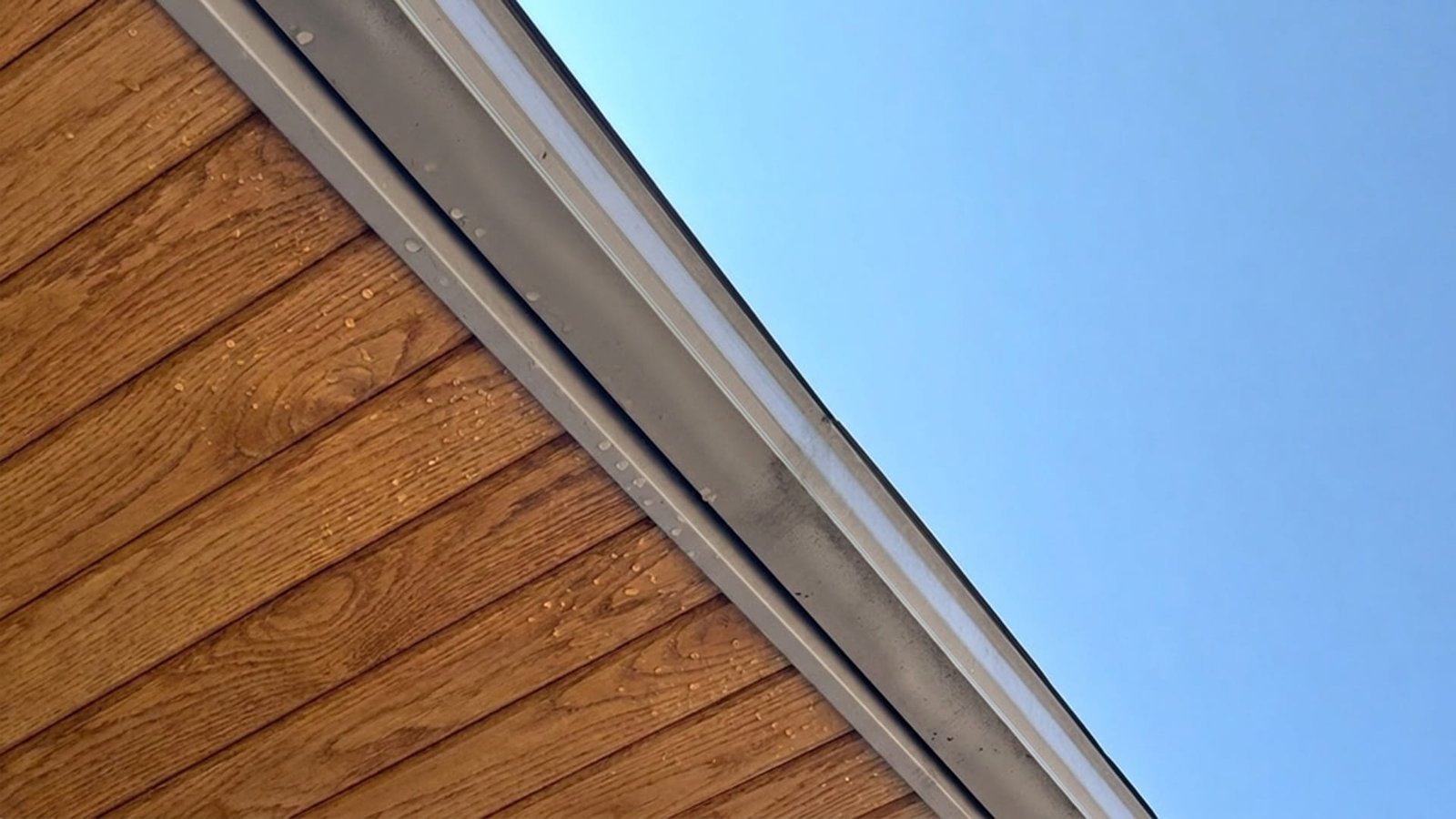 Close-up of a wood-panelled roofline edge with a metal gutter and drip edge against a clear blue sky