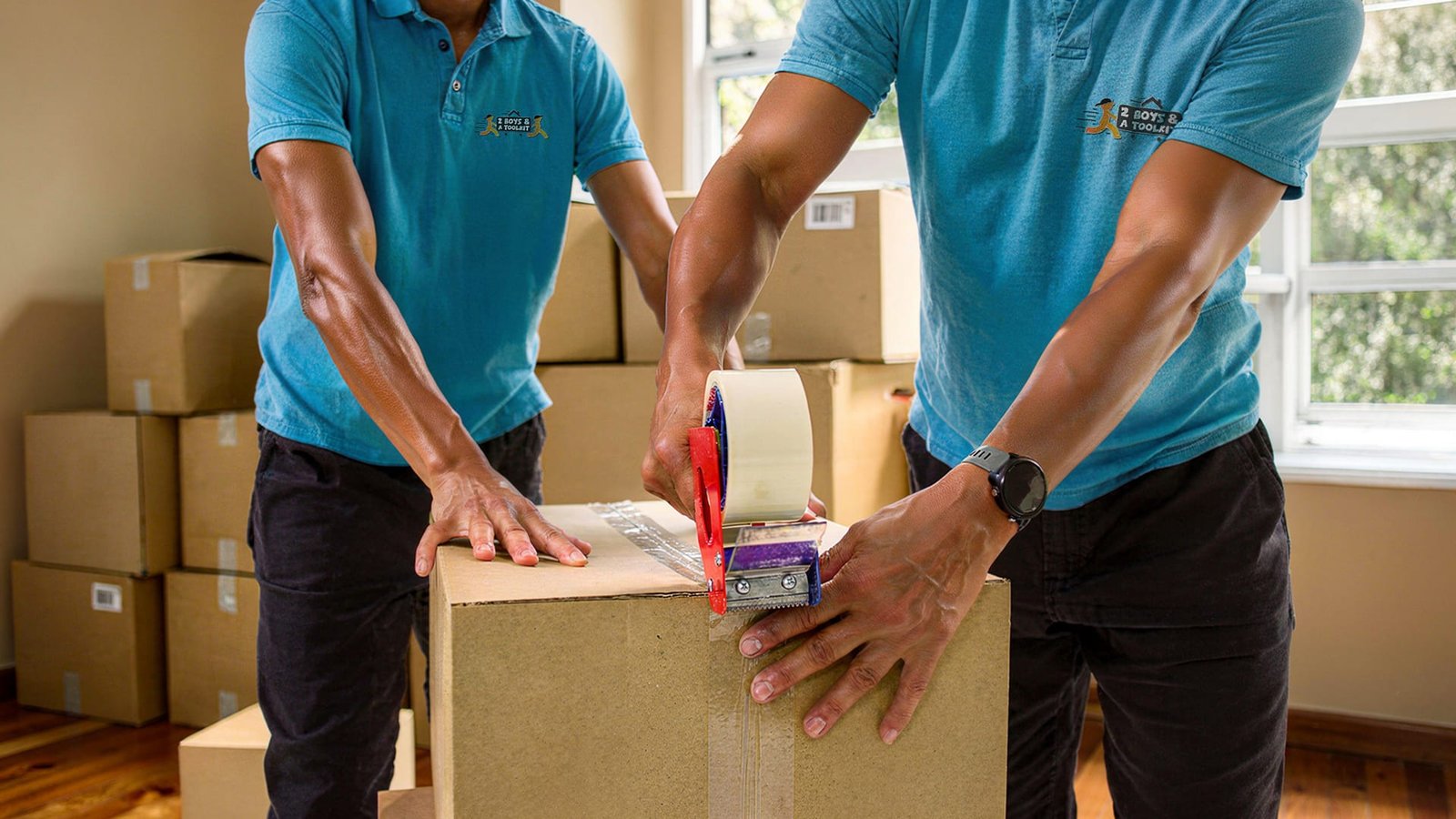 Two 2 Boys and a Toolkit movers in teal shirts taping a moving box, surrounded by stacked cardboard boxes