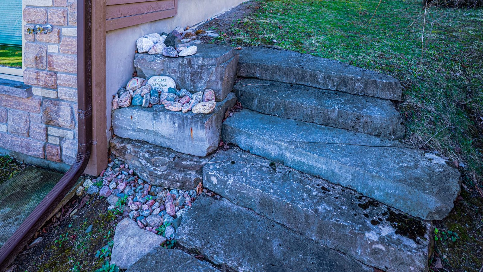 Stone steps beside a house, with a painted rock reading "Mom's Garden" among a collection of decorative rocks