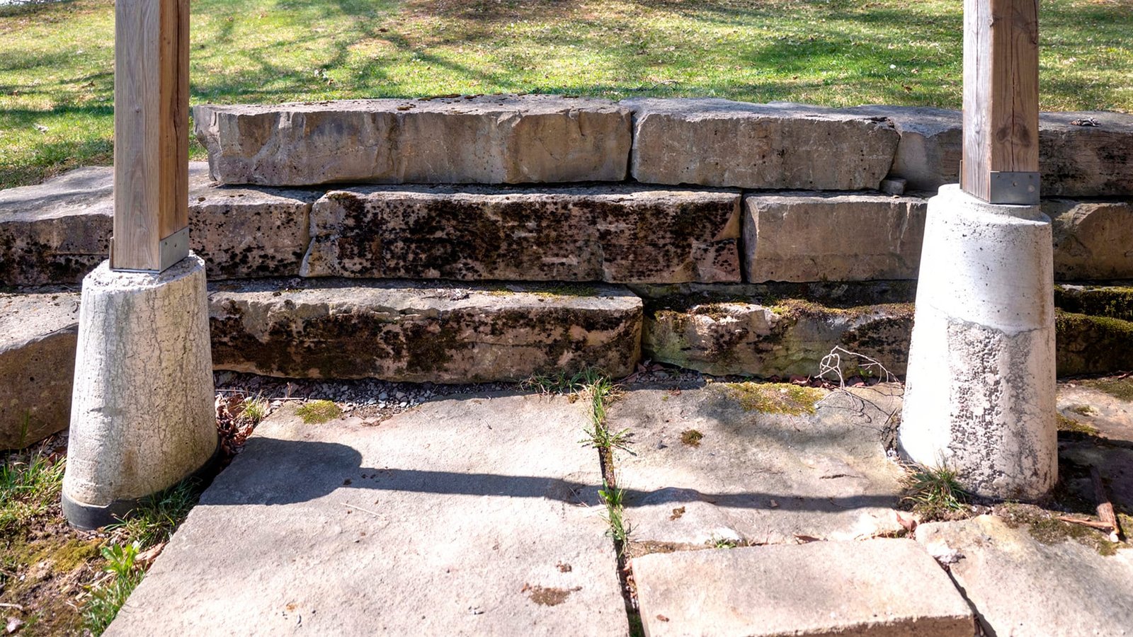 Weathered stone steps with moss between wooden posts on concrete footings, leading to a grassy lawn