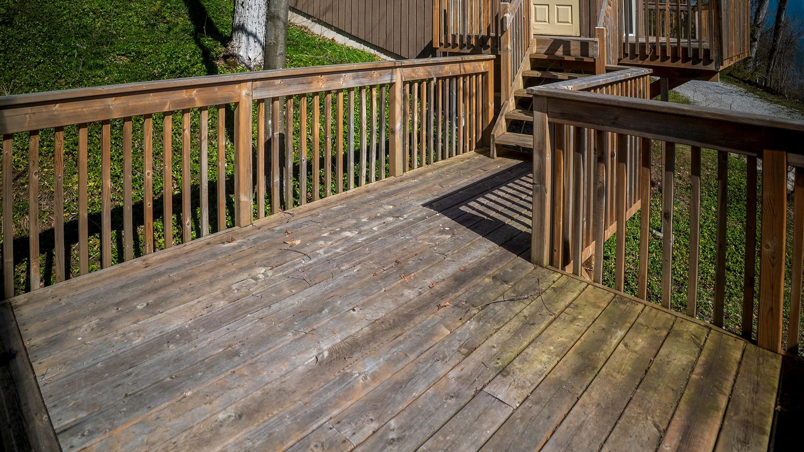 Weathered wooden deck with railings and stairs leading to the house entrance, surrounded by grassy hillside