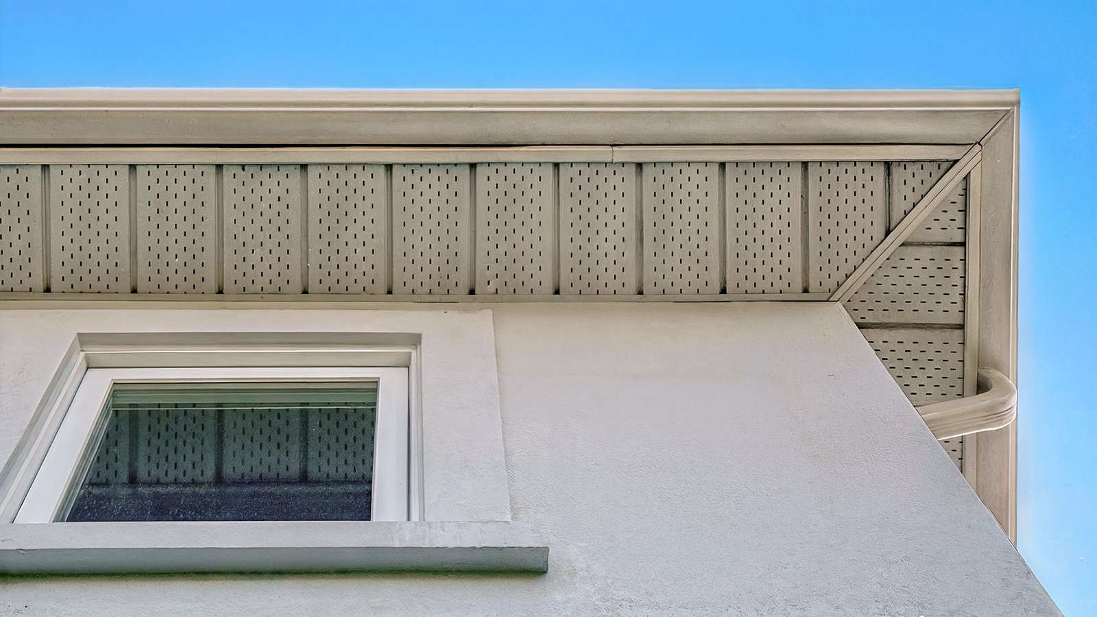 Upward view of a stucco house corner with vented eaves, wooden fascia, and a window against a blue sky