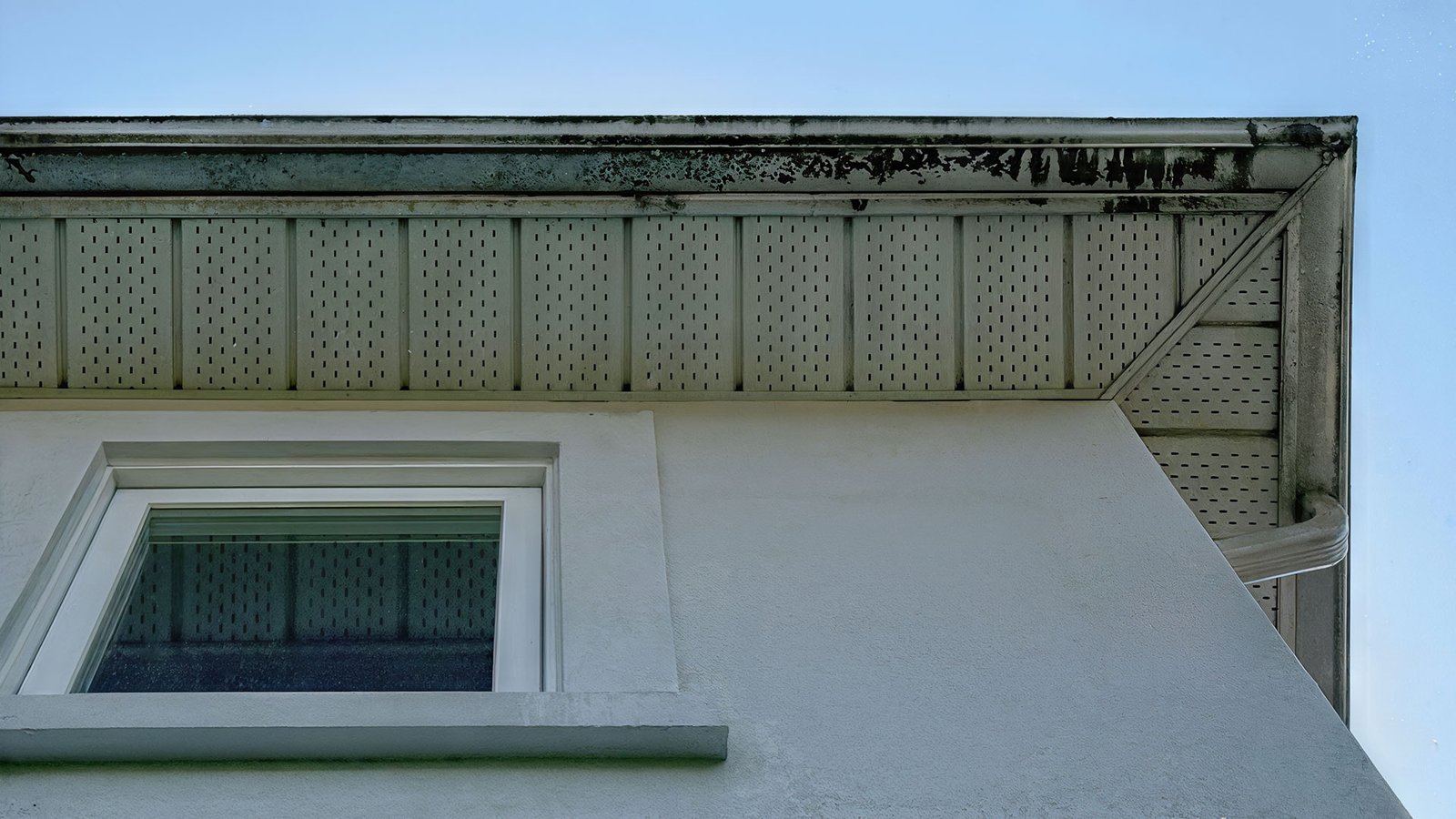 Upward view of a stucco building corner with a perforated wooden soffit and weathered eaves against a blue sky