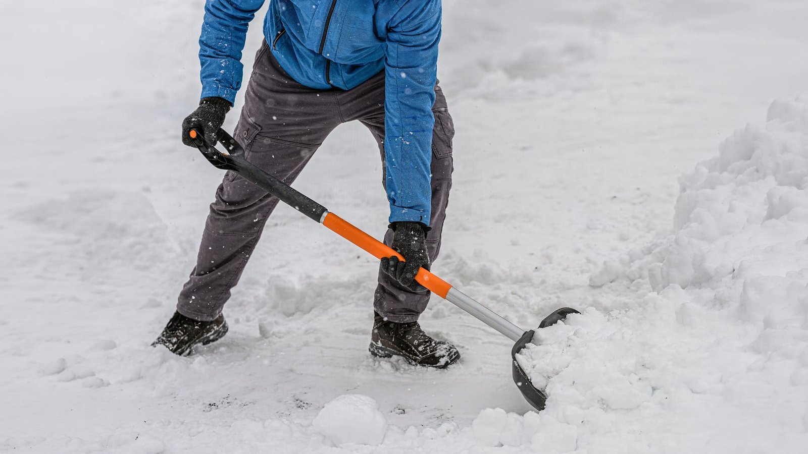 Person in a blue jacket and grey pants shovelling snow with an orange-handled shovel